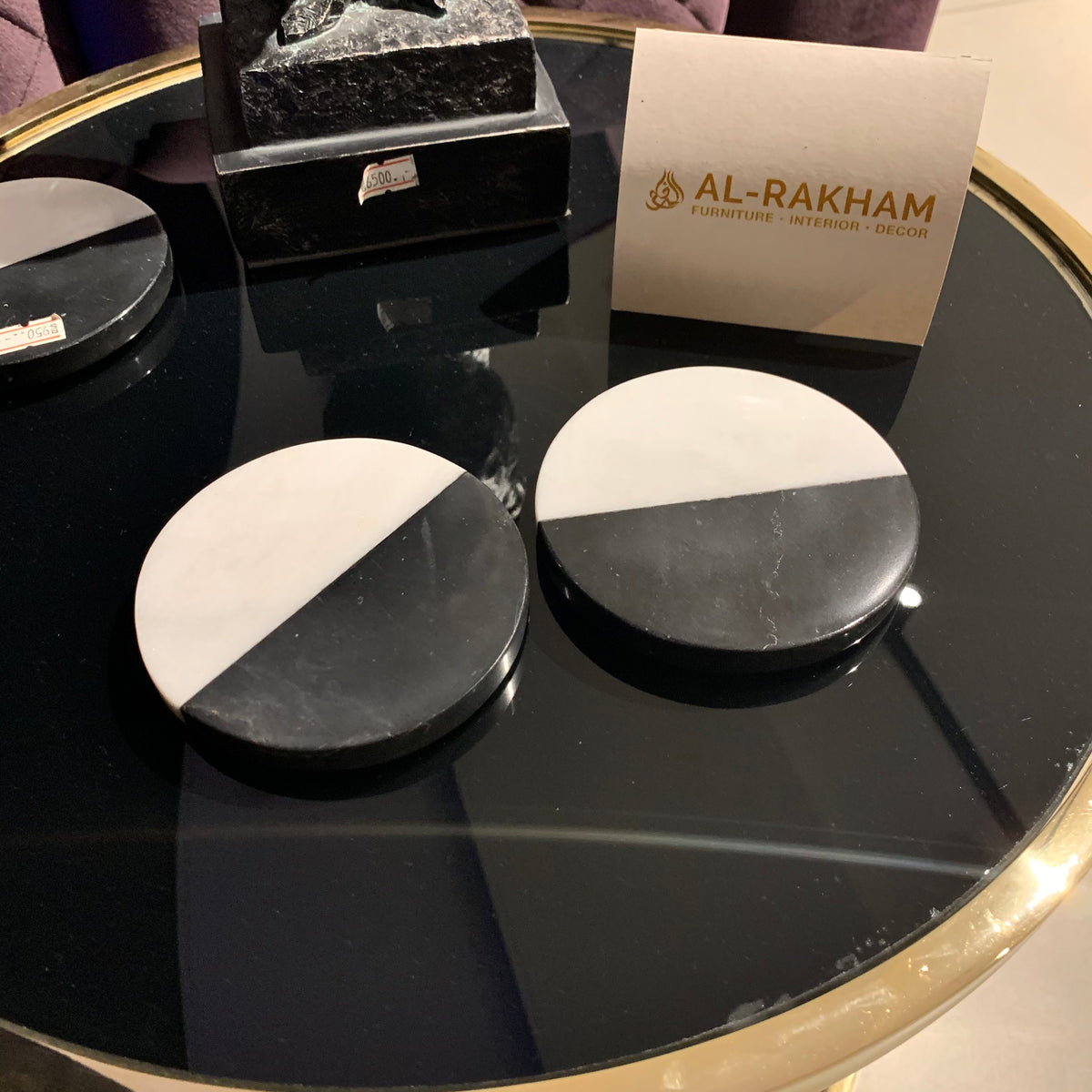 Black and white coasters on a reflective surface with 'Al-Rakham' branding in the background.