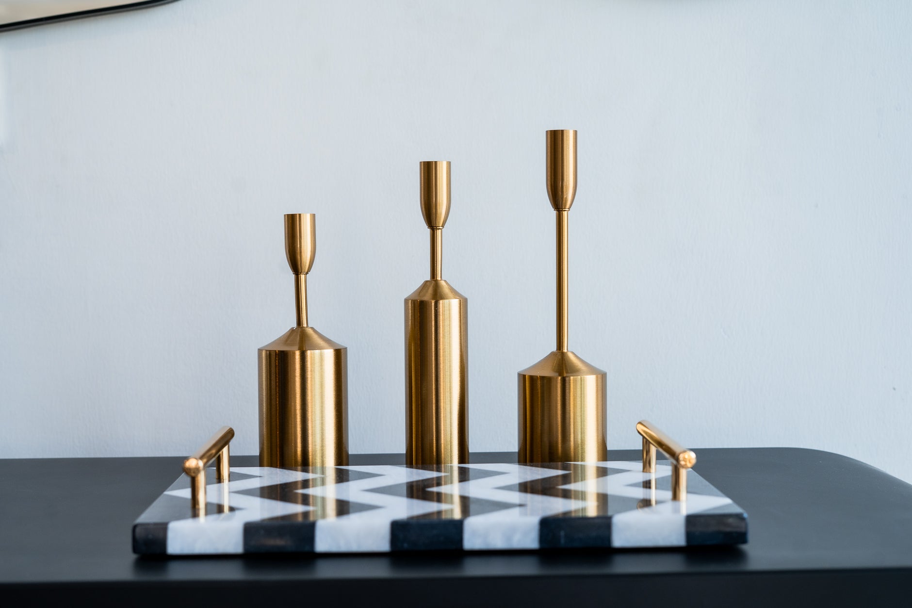 Set of gold candle holders on a black and white patterned tray against a light gray wall.