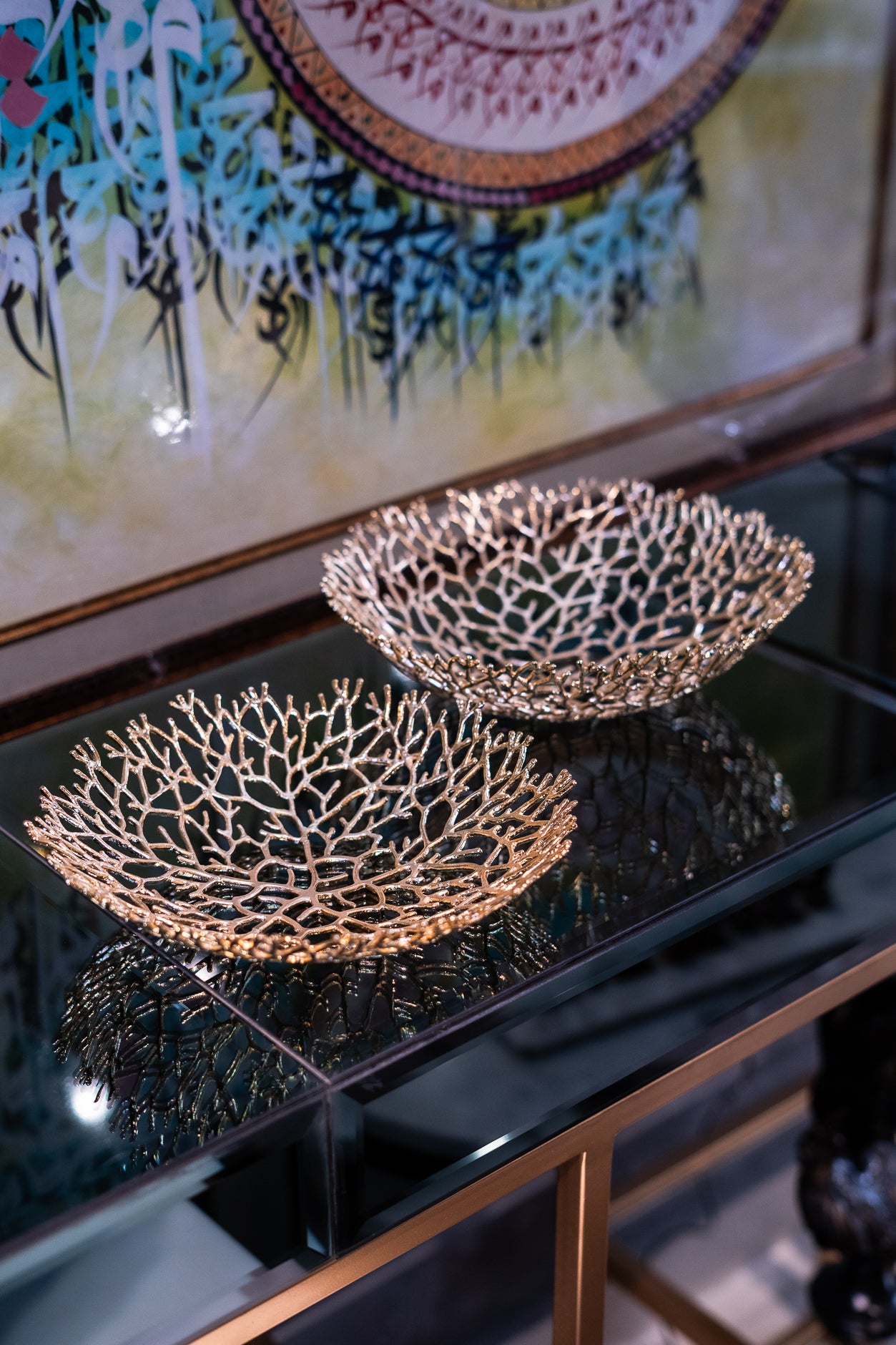 Two decorative bowls with a coral design on a reflective surface, with a colorful abstract painting in the background.