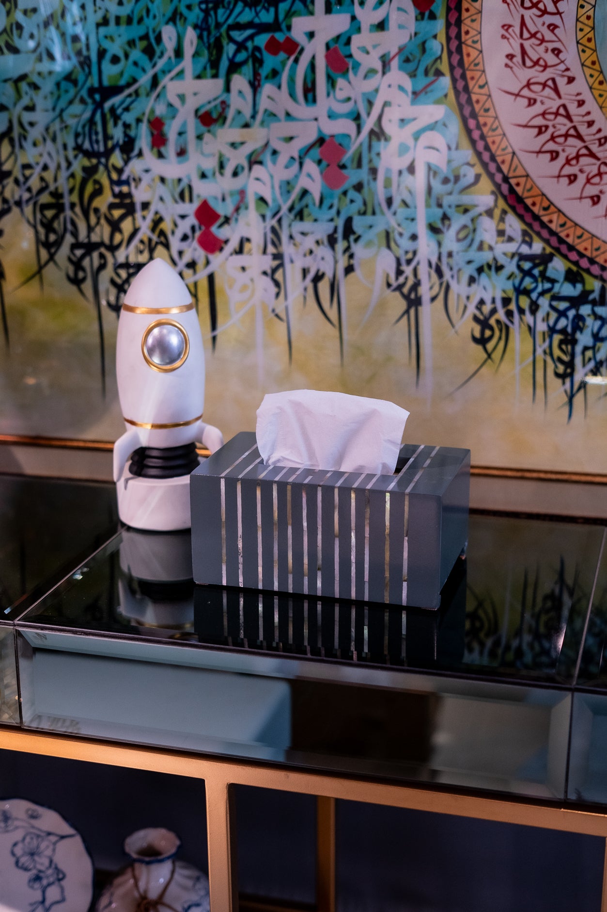 Tissue box with a decorative robot figure on a reflective surface with a colorful abstract painting in the background.