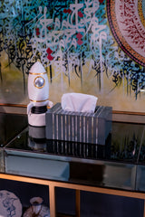 Tissue box with a decorative robot figure on a reflective surface with a colorful abstract painting in the background.